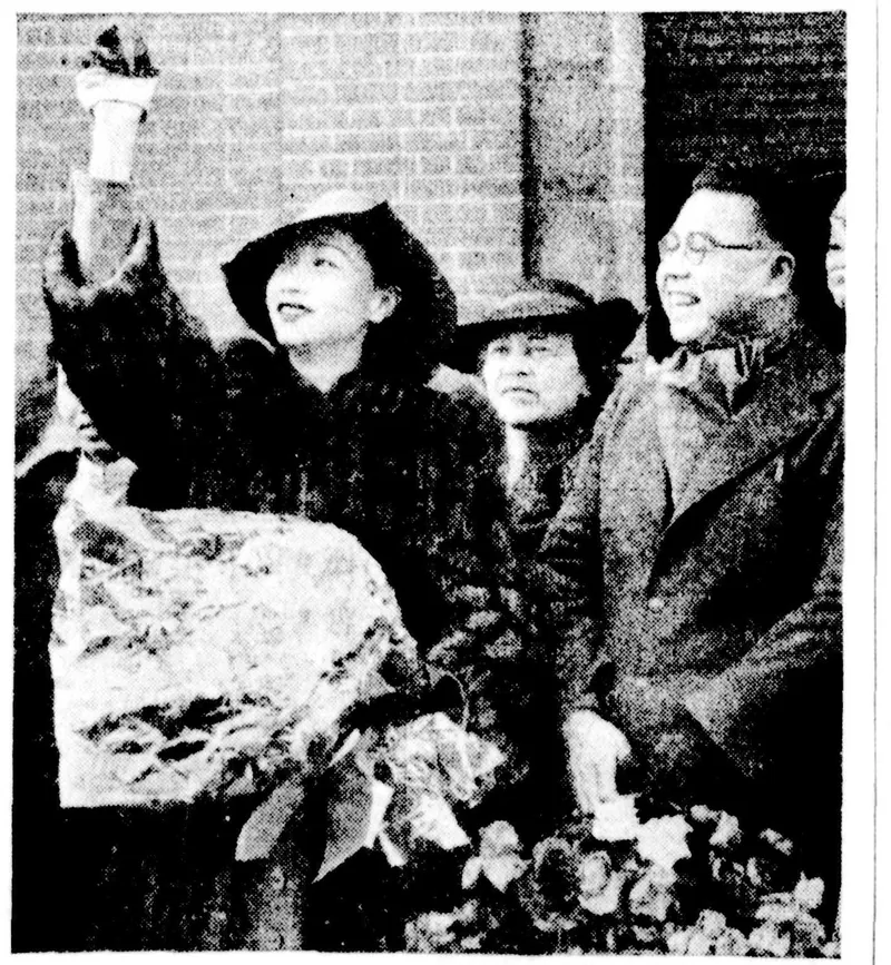 Anna May Wong in a wide hat and fur, waving, flowers in her arms, among a crowd at a station.