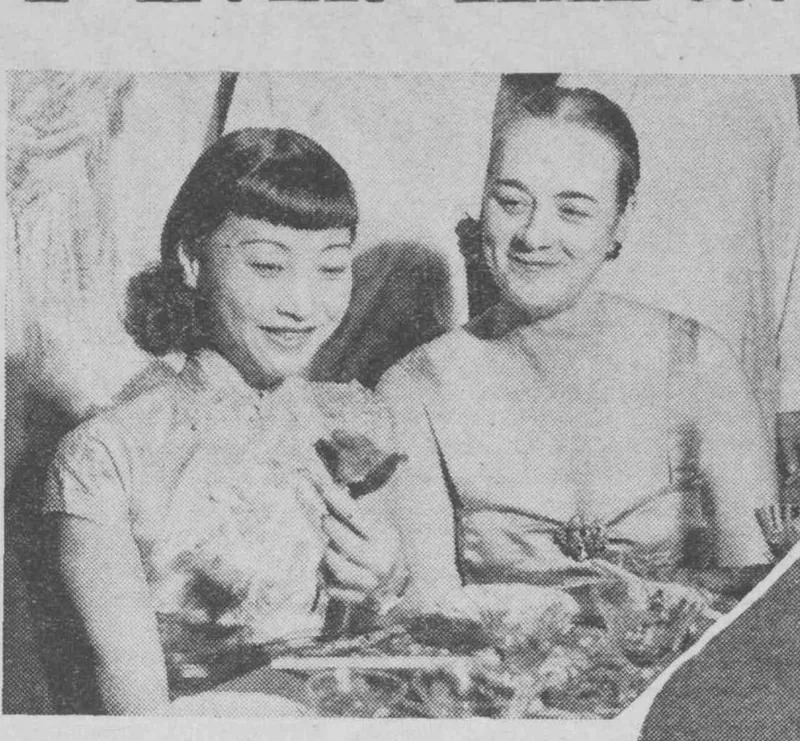 Anna May Wong at a dinner table holding a rose, beside a woman in an evening dress.